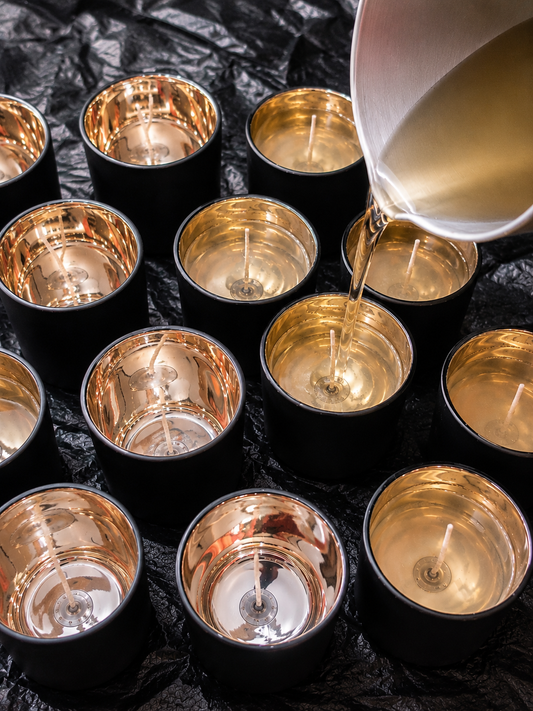 Luxury candle being hand-poured into a matte black jar with gold interior, surrounded by fragrance oils and candle-making tools in soft lighting.